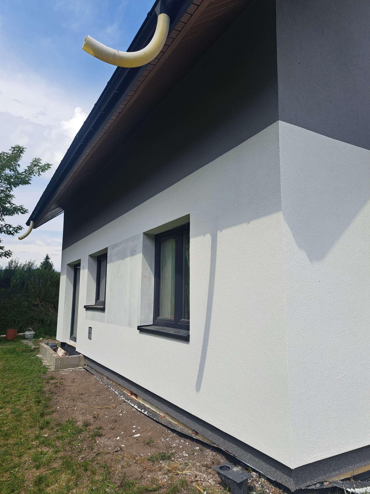 Dwukolorowa elewacja domu jednorodzinnego z widocznym fragmentem ocieplenia przy gruncie i żółtą rurą spustową. Świeżo położony tynk strukturalny.
