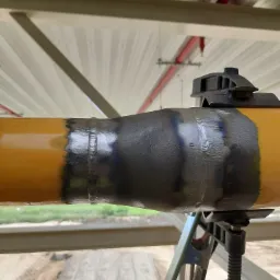 Żółta rura z widocznym spawem, podpórką i oznaczeniami norm, umieszczona w konstrukcji stalowej.