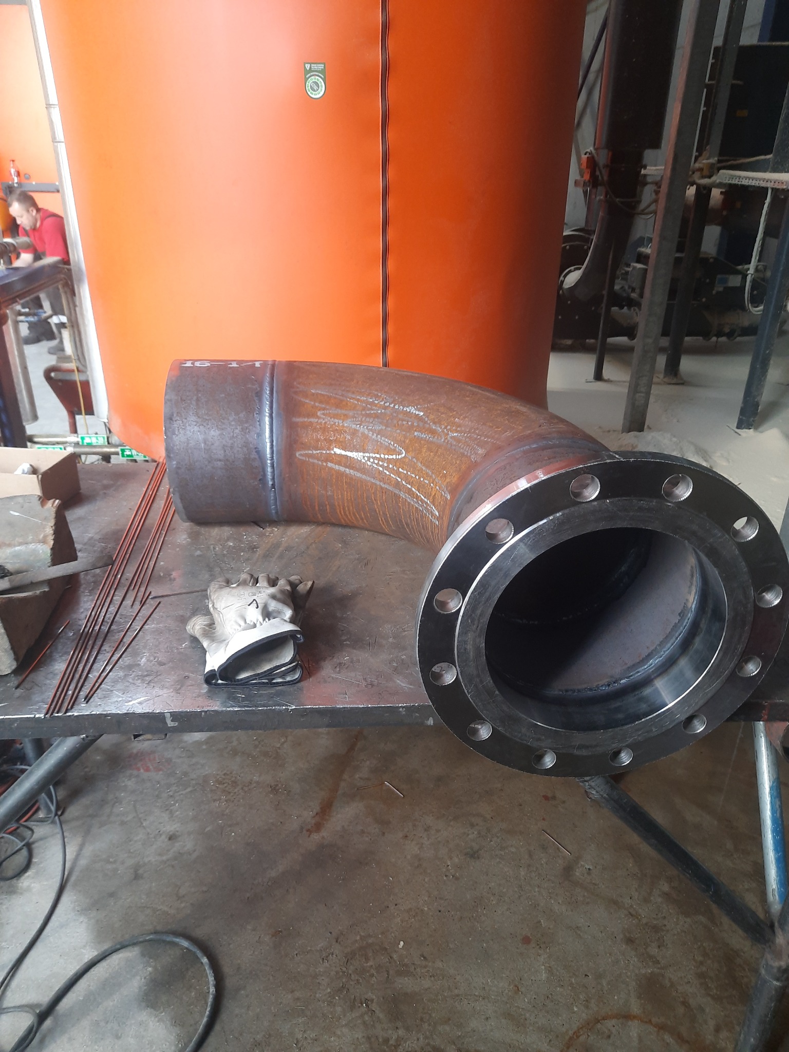 Spawany kolanko rury z kołnierzem na metalowym stole warsztatowym, obok elektrody spawalnicze i rękawice robocze, w tle pomarańczowy zbiornik i spawacz przy pracy.