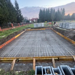 M&S PARTNERS MYKHAILO RIAZANTSEV - Zbrojenie fundamentu domu jednorodzinnego w szalunku, widok z góry. Na drugim planie cegły, bloczki i pracownicy budowlani. W tle dom i drzewa.