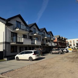 M&S PARTNERS MYKHAILO RIAZANTSEV - Nowoczesne, szeregowe domy z balkonami i szklanymi balustradami w słoneczny dzień. Biały samochód zaparkowany przed budynkiem. Częściowo widoczny budynek w trakcie budowy w tle.