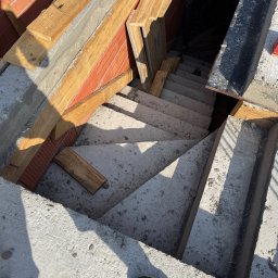 M&S PARTNERS MYKHAILO RIAZANTSEV - Betonowe schody w trakcie budowy, widok z góry. Szalunki z drewna i cegieł. Cień osoby w rogu kadru. Surowy stan, etap prac budowlanych.