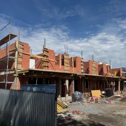 M&S PARTNERS MYKHAILO RIAZANTSEV - Wznoszenie murów z czerwonej cegły na budowie osiedla, widoczne rusztowania, szalunki i wystające pręty zbrojeniowe. W tle błękitne niebo z chmurami.