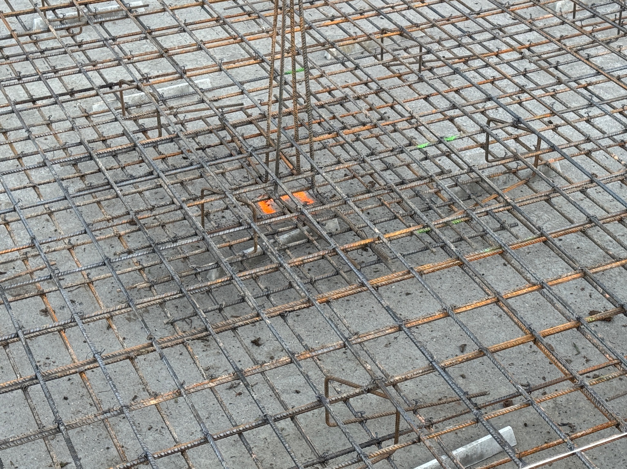 Zbrojenie fundamentu z wystającymi prętami pionowymi, tworzące siatkę na szarym betonie. Widoczne pomarańczowe elementy dystansowe.