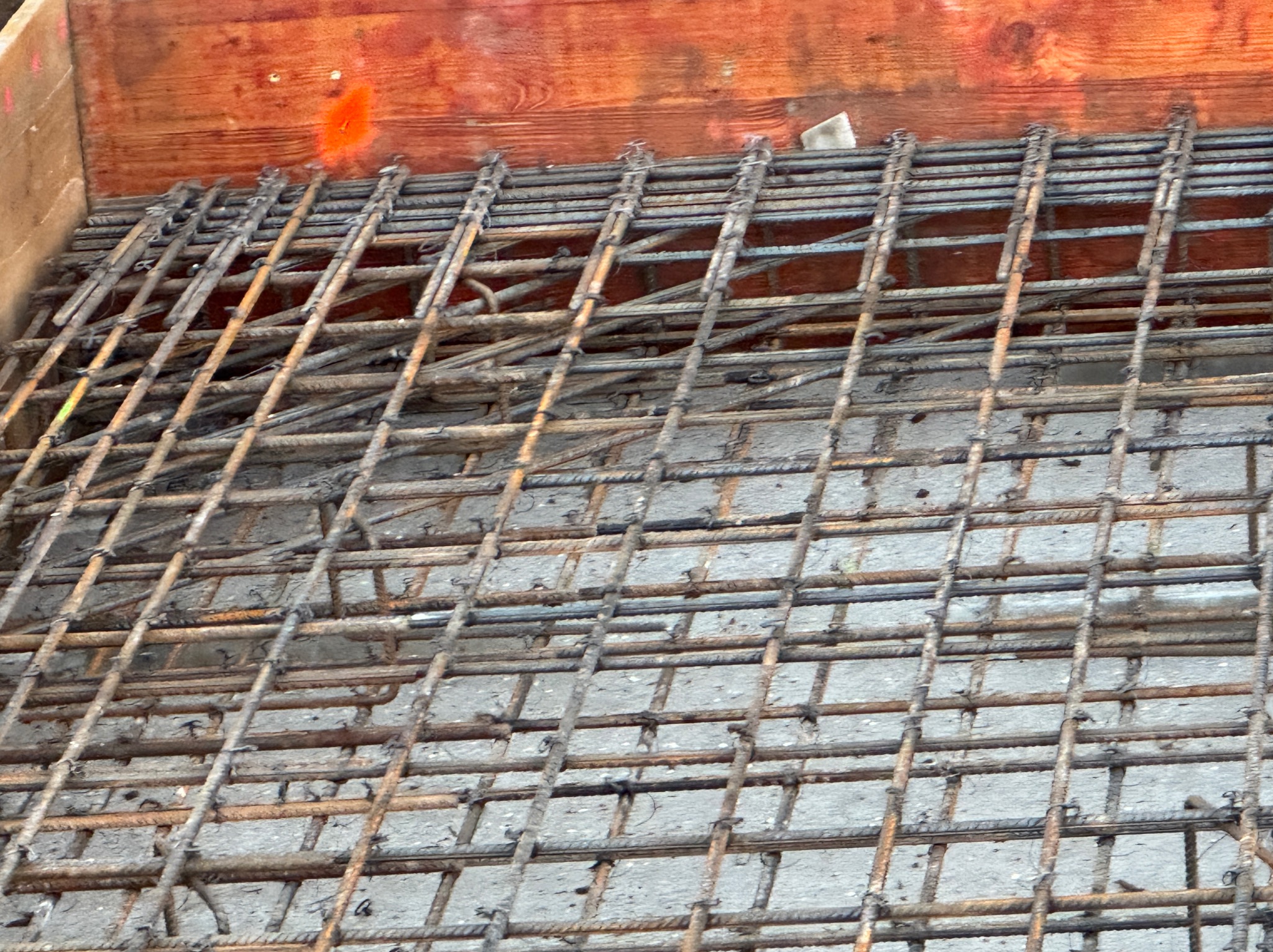 Zbrojenie fundamentu w drewnianym szalunku, widok z góry. Stalowe pręty tworzą regularną siatkę, przygotowaną do zalania betonem. Detal budowy.