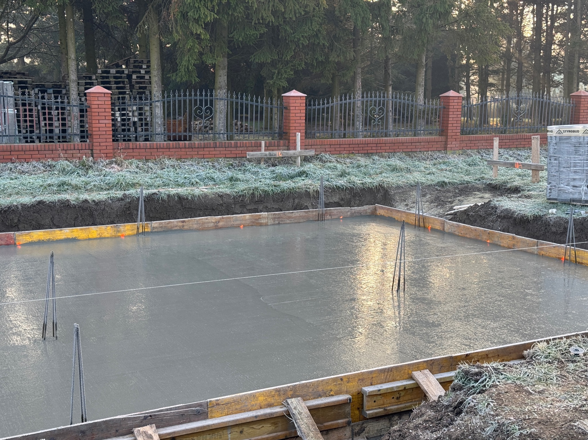Zalany fundament pod budowę domu, widoczne szalunki i wystające pręty zbrojeniowe. W tle ogrodzenie z cegły i metaloplastyki, drzewa.