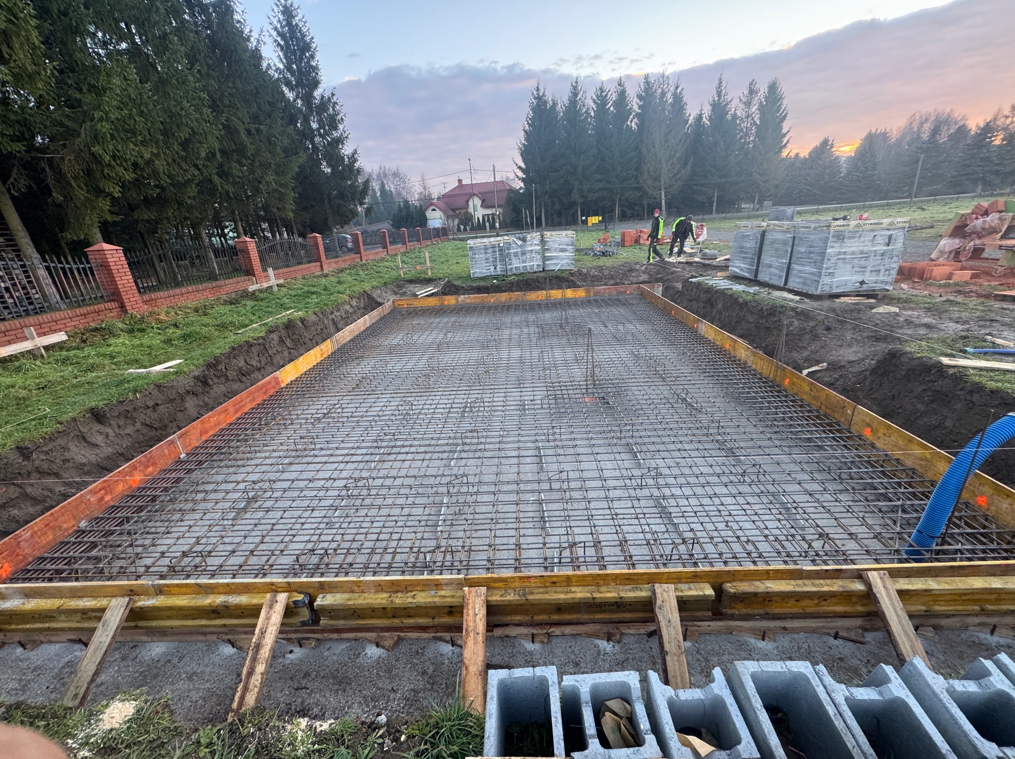 Zbrojenie fundamentu domu jednorodzinnego w szalunku, widok z góry. Na drugim planie cegły, bloczki i pracownicy budowlani. W tle dom i drzewa.