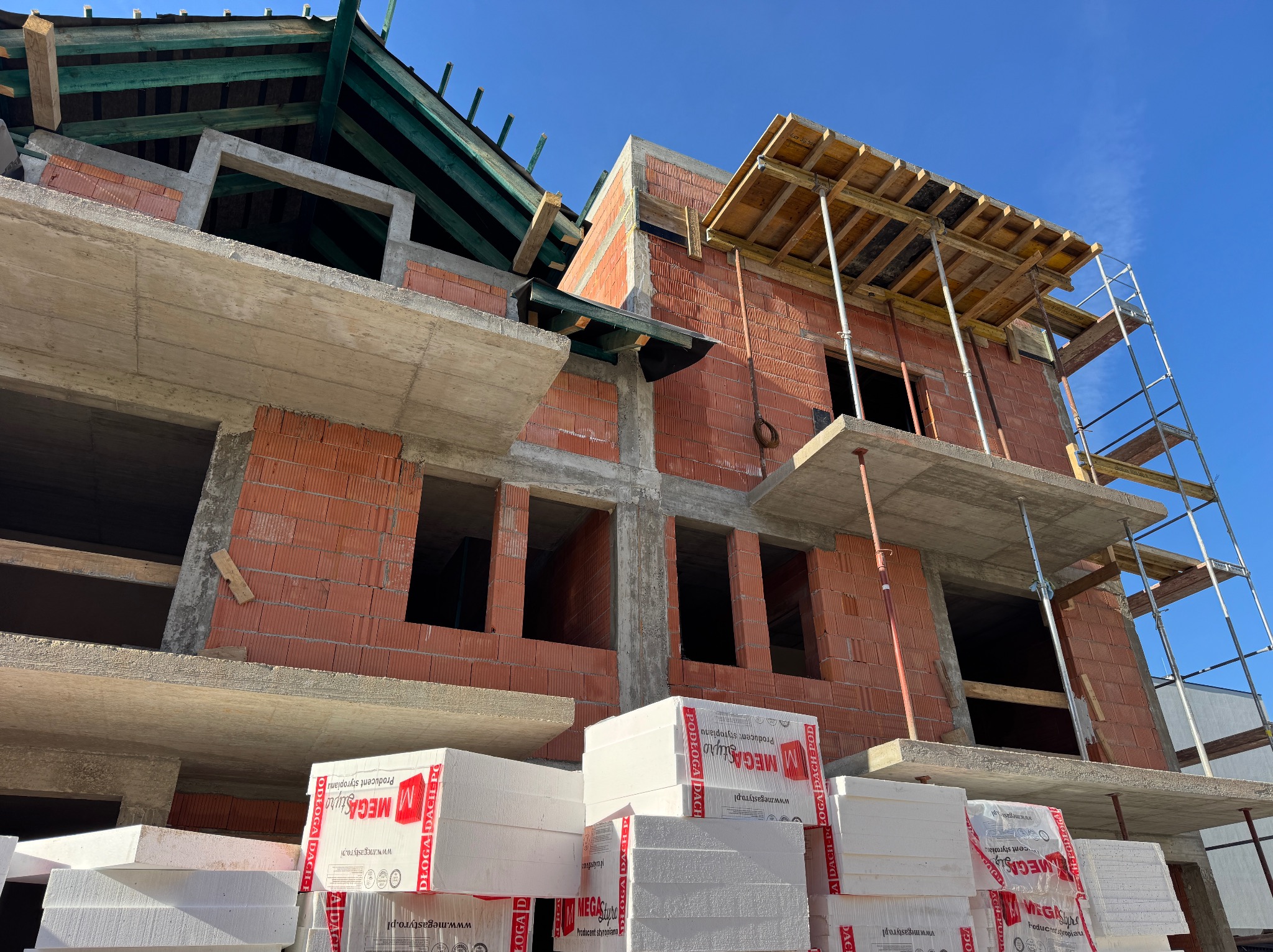 Surowy stan domu murowanego z czerwonej cegły, widoczne szalunki stropowe i rusztowanie. Na pierwszym planie paczki styropianu.