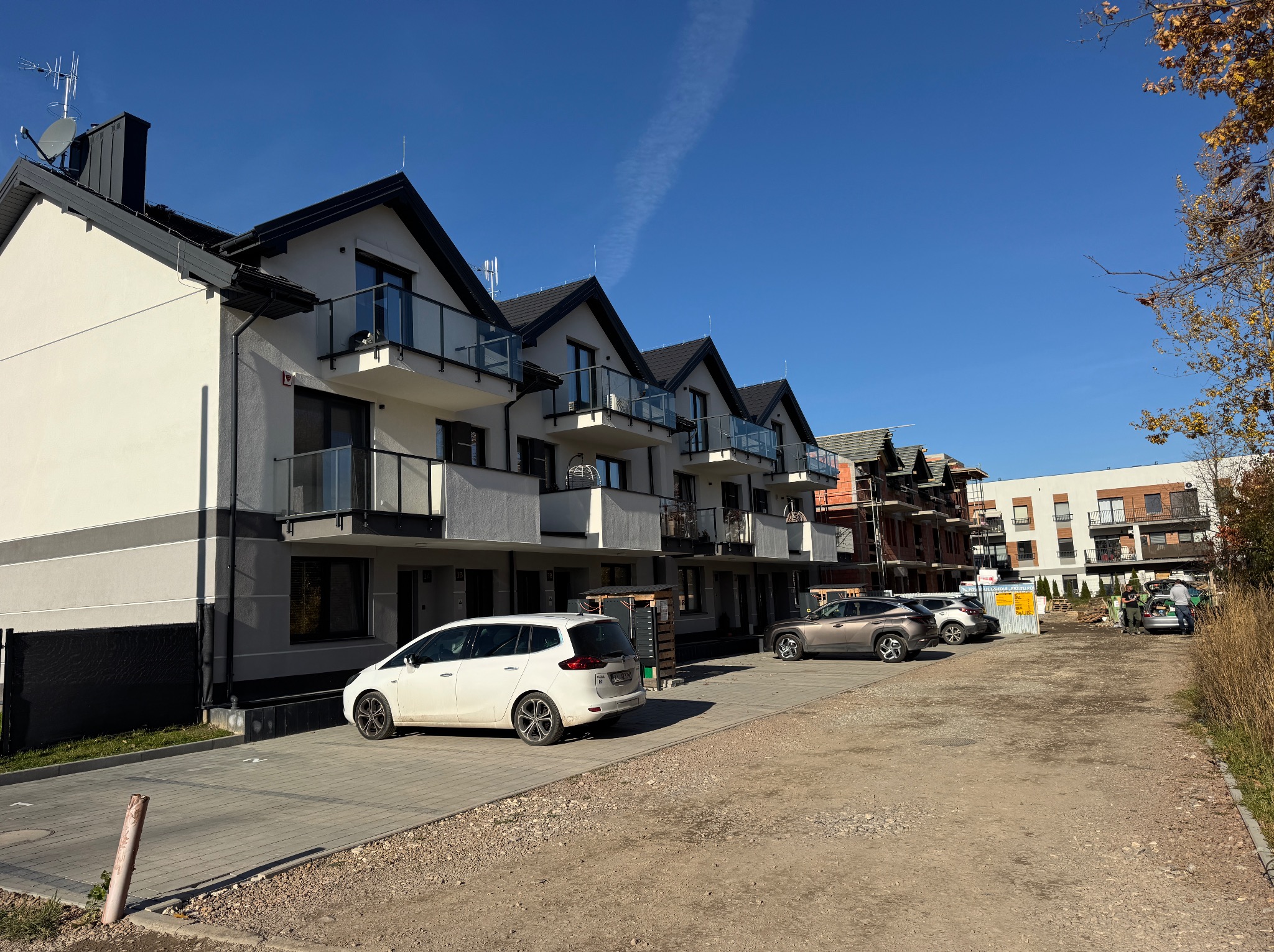 Nowoczesne, szeregowe domy z balkonami i szklanymi balustradami w słoneczny dzień. Biały samochód zaparkowany przed budynkiem. Częściowo widoczny budynek w trakcie budowy w tle.