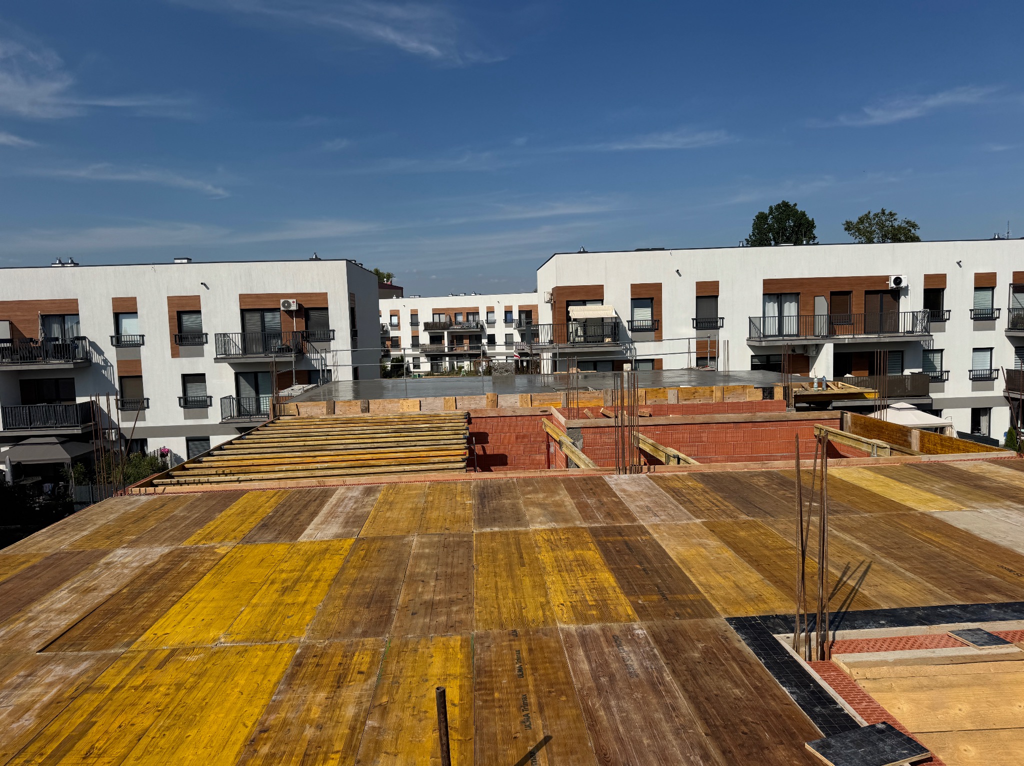 Budowa wielorodzinnego domu: szalunek stropu z żółtych płyt, zbrojenie, w tle białe budynki z balkonami i błękitne niebo. Widok z góry na plac budowy.