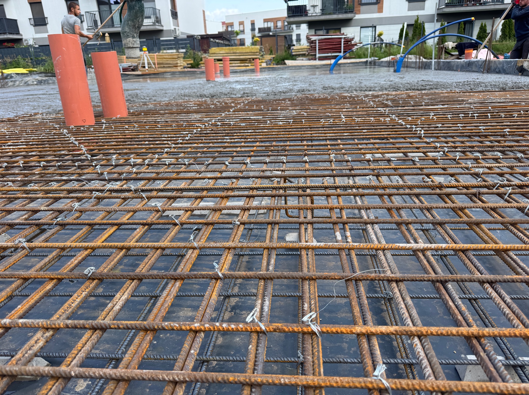 Zbrojenie fundamentów z widoczną siatką stalową i wylanym betonem w tle, na budowie nowego budynku mieszkalnego. Widoczne pomarańczowe rury i pracownik.