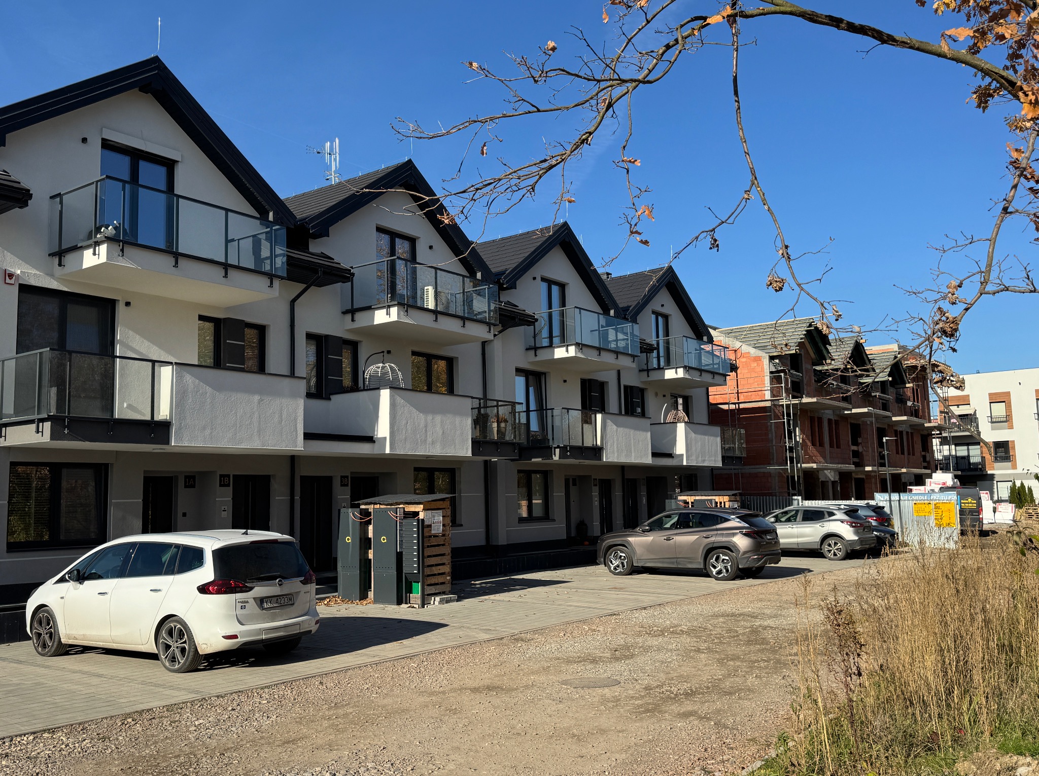 Nowoczesne domy szeregowe z balkonami i szklanymi balustradami, zaparkowane samochody, budowa w tle, słoneczny dzień, jesienne gałęzie drzewa w kadrze.