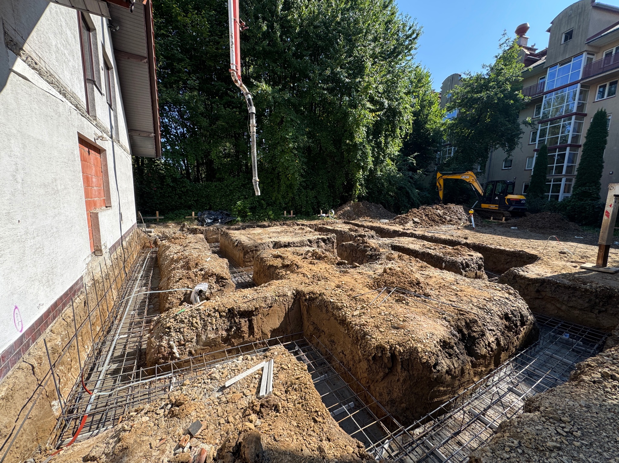 Wykopy pod fundamenty zbrojone stalą, widoczny budynek w tle i ramię pompy do betonu. Prace ziemne i przygotowanie do wylewania fundamentów.
