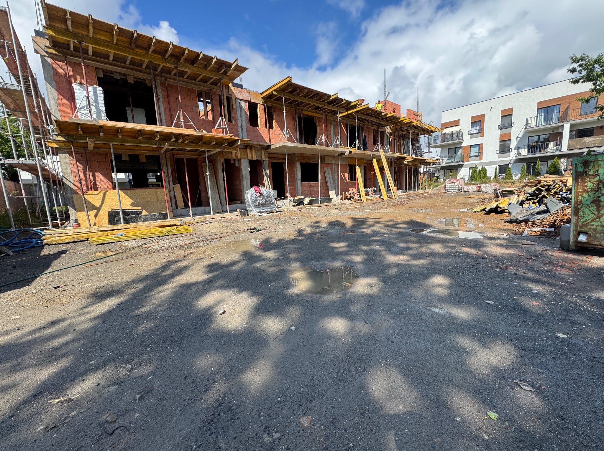 Wznoszenie dwukondygnacyjnego domu murowanego z czerwonej cegły, z widocznym szalunkiem stropu i elementami rusztowań. W tle budynek mieszkalny i błękitne niebo.