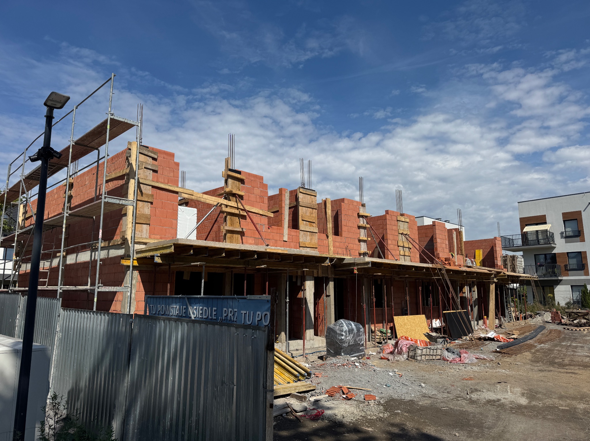 Wznoszenie murów z czerwonej cegły na budowie osiedla, widoczne rusztowania, szalunki i wystające pręty zbrojeniowe. W tle błękitne niebo z chmurami.