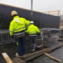 Dwóch zbrojarzy w jaskrawych kurtkach przy pracy z prętami zbrojeniowymi na tle szalunku ściennego ULMA, widok z tyłu.