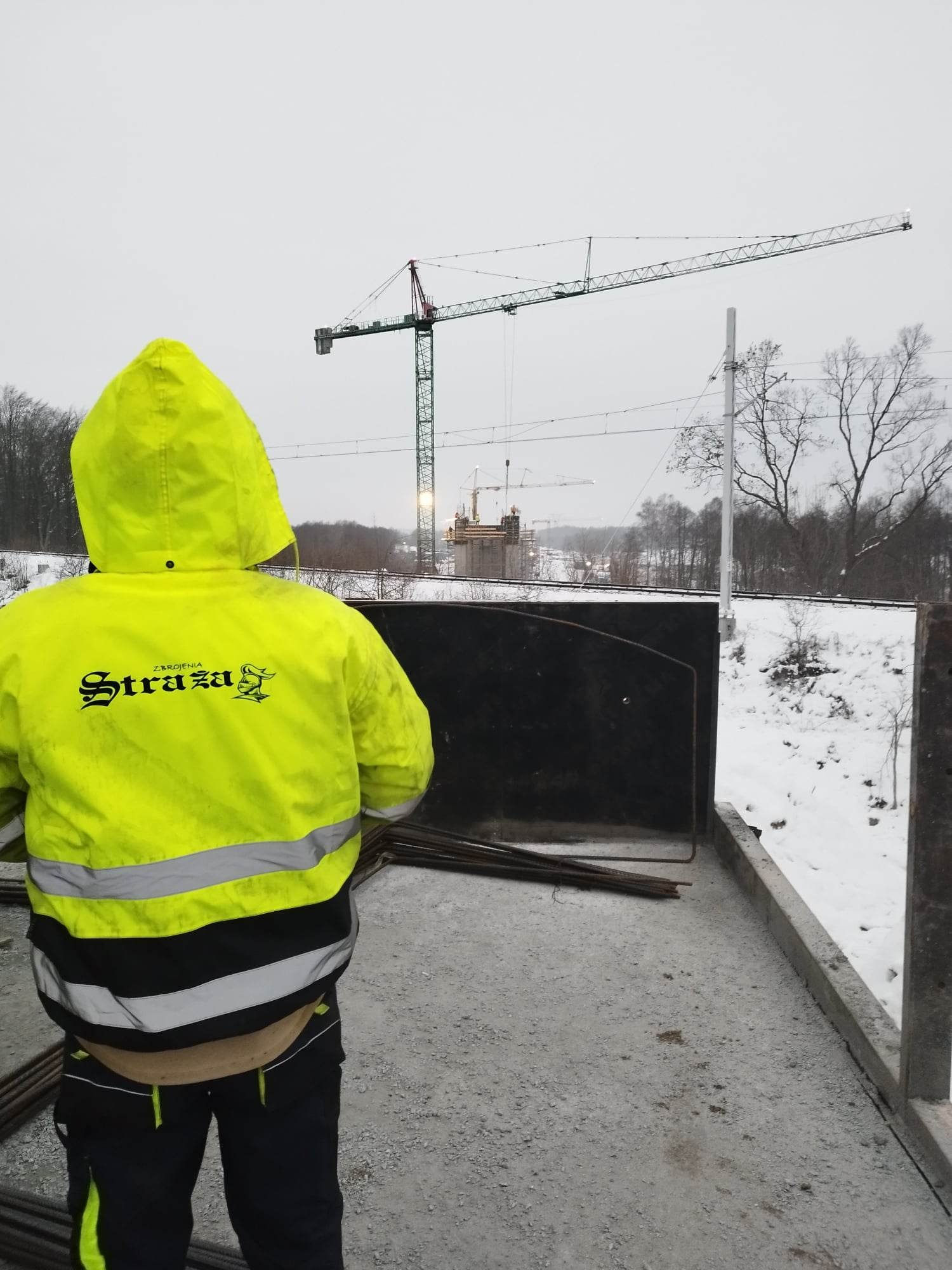 Pracownik w żółtej kurtce z napisem 'Straza' na budowie budynku żelbetowego, widoczne zbrojenia i żuraw budowlany w tle, zimowa sceneria.