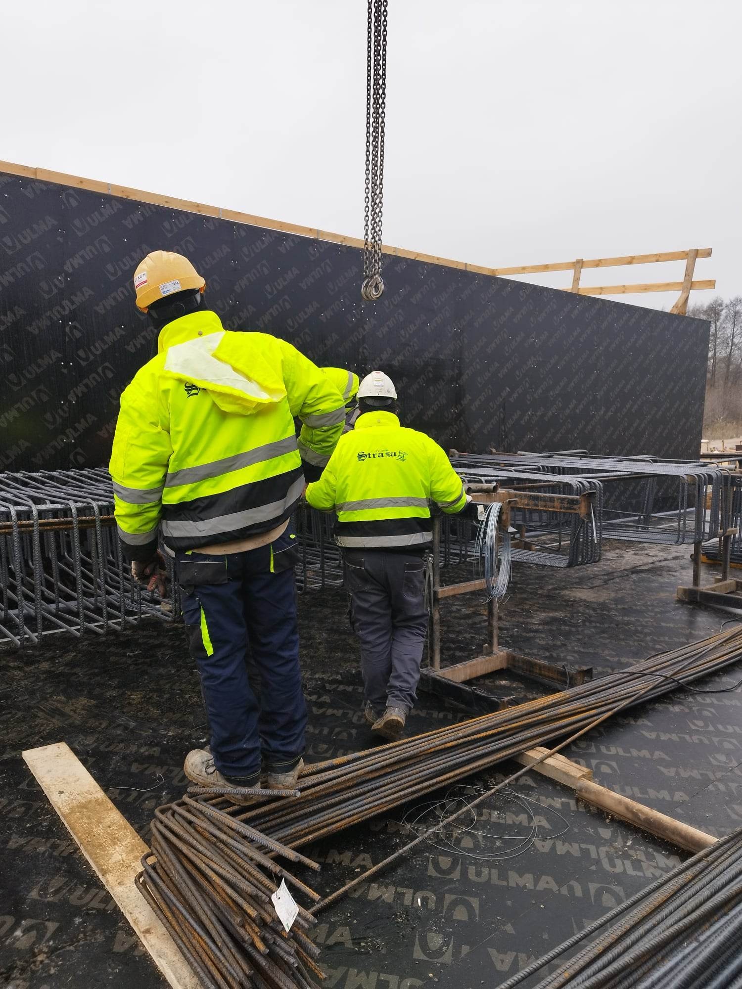 Dwóch zbrojarzy w jaskrawych kurtkach przy pracy z prętami zbrojeniowymi na tle szalunku ściennego ULMA, widok z tyłu.