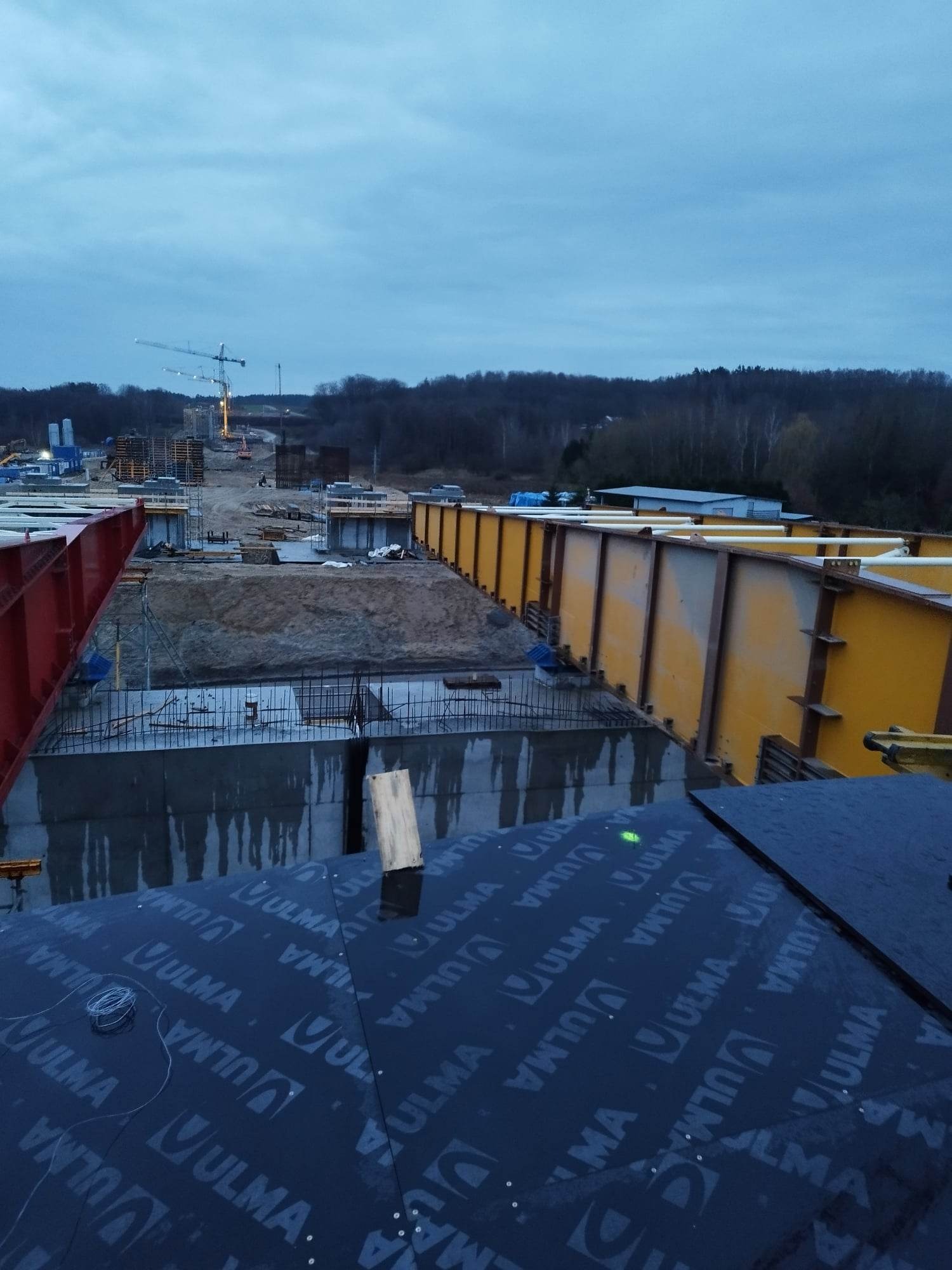 Wiązanie zbrojenia na budowie mostu, widoczne szalunki, dźwigi w tle i materiał izolacyjny z logo ULMA na pierwszym planie.