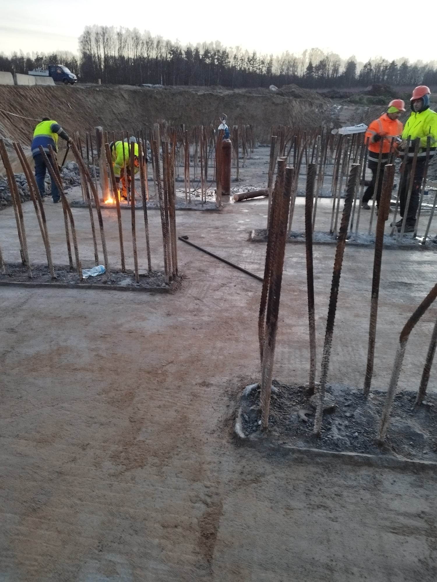 Prace zbrojarskie na fundamencie budynku: spawanie prętów zbrojeniowych przez pracownika w żółtej kamizelce, widoczni inni pracownicy w odblaskowych ubraniach i butla z gazem w tle.