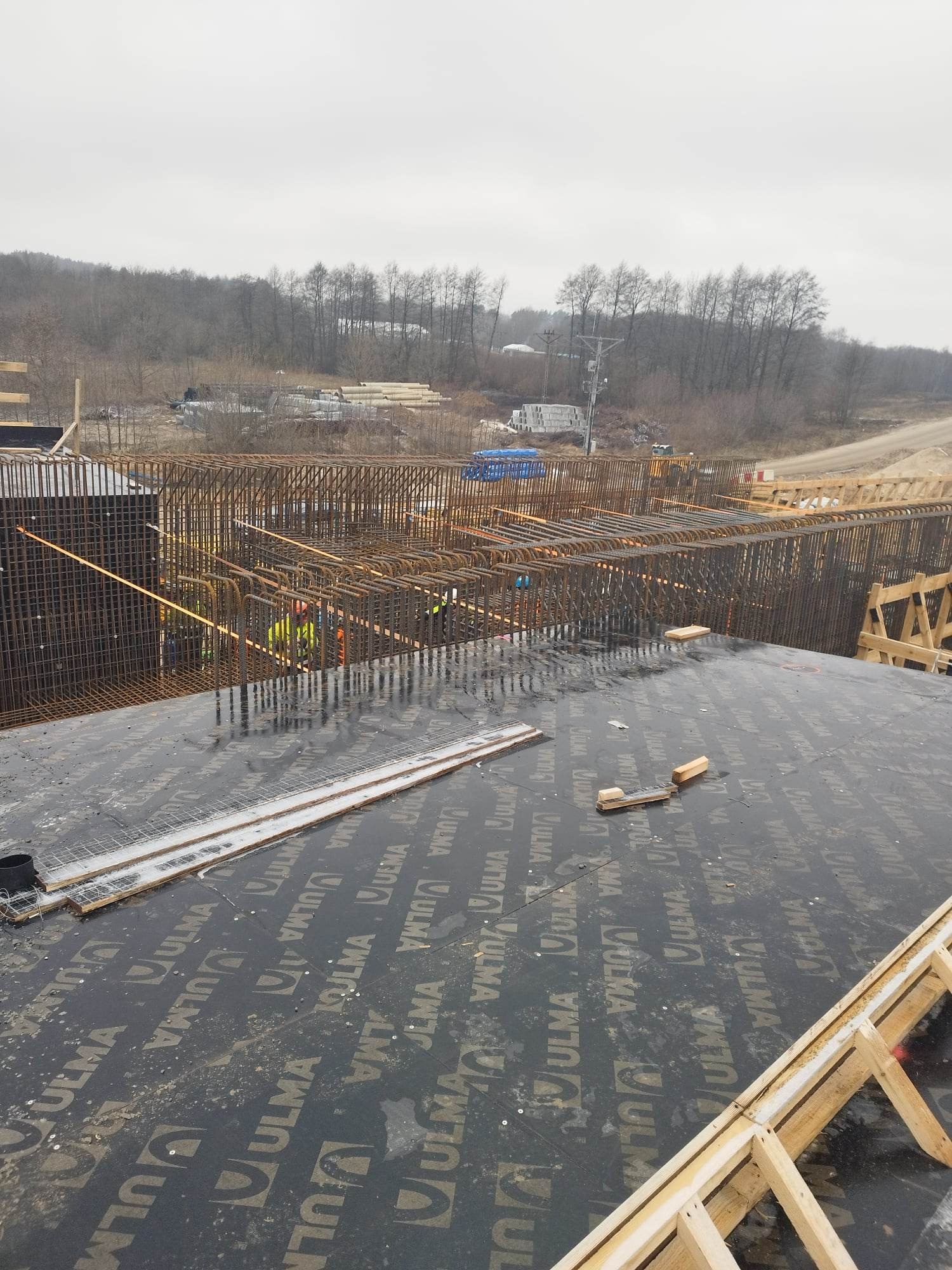 Wiązanie zbrojenia na budowie w Kamiennej Górze, widok z góry na mokrą, pokrytą deskami szalunkowymi powierzchnię, w tle widoczni pracownicy w odblaskowych kamizelkach i stalowe pręty zbrojeniowe.