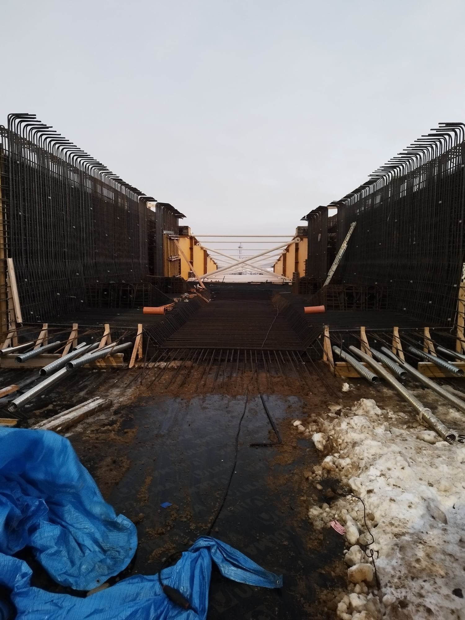 Konstrukcja zbrojeniowa pod budowę mostu w trakcie montażu, widoczne stalowe pręty zagięte w charakterystyczny sposób, szalunki, elementy rusztowania i pozostałości śniegu na ziemi.