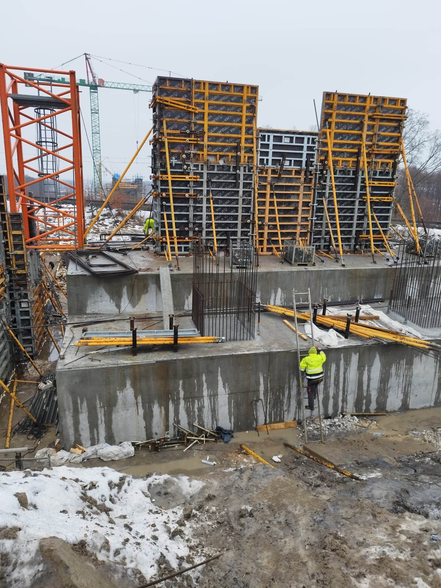 Wznoszenie żelbetowej konstrukcji: pracownik w żółtej kurtce wchodzi po drabinie na szalunek fundamentu, widoczne zbrojenia, dźwig w tle i błotniste podłoże z resztkami śniegu.