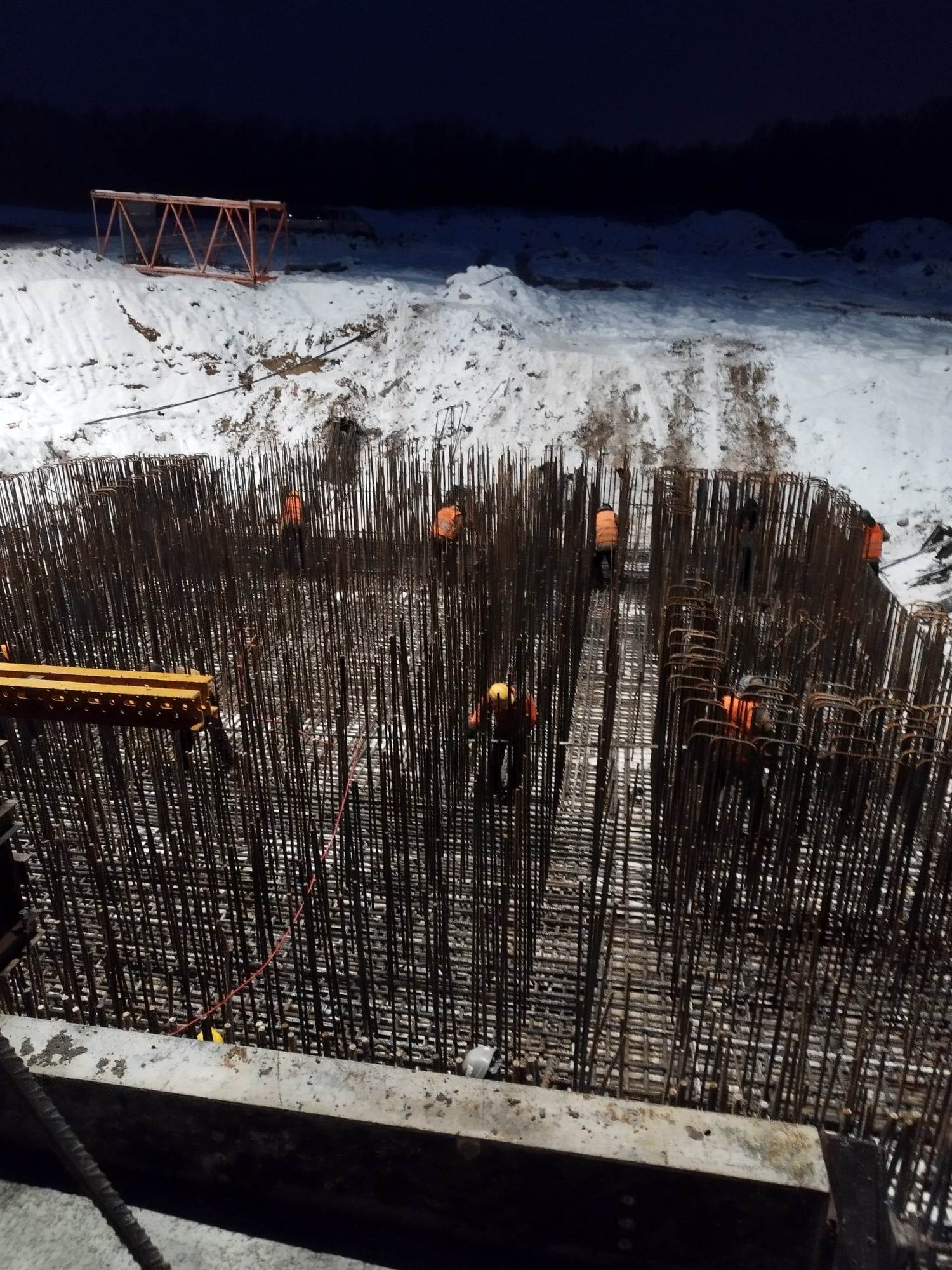 Zimowa budowa: kilku pracowników w pomarańczowych kamizelkach układa gęstą siatkę z prętów zbrojeniowych w wykopie zasypanym śniegiem, oświetlonym sztucznym światłem.