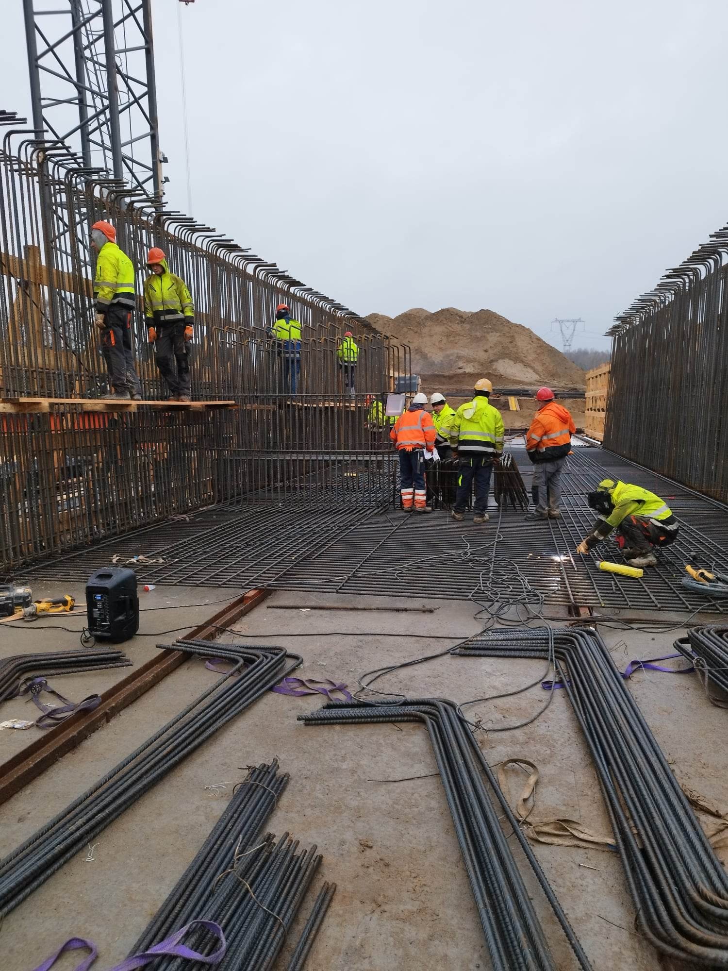 Konstrukcja zbrojeniowa mostu w trakcie budowy; widoczni pracownicy w odblaskowych ubraniach, spawarka w użyciu, przygotowane pręty zbrojeniowe na ziemi, w tle hałda ziemi i słup wysokiego napięcia.