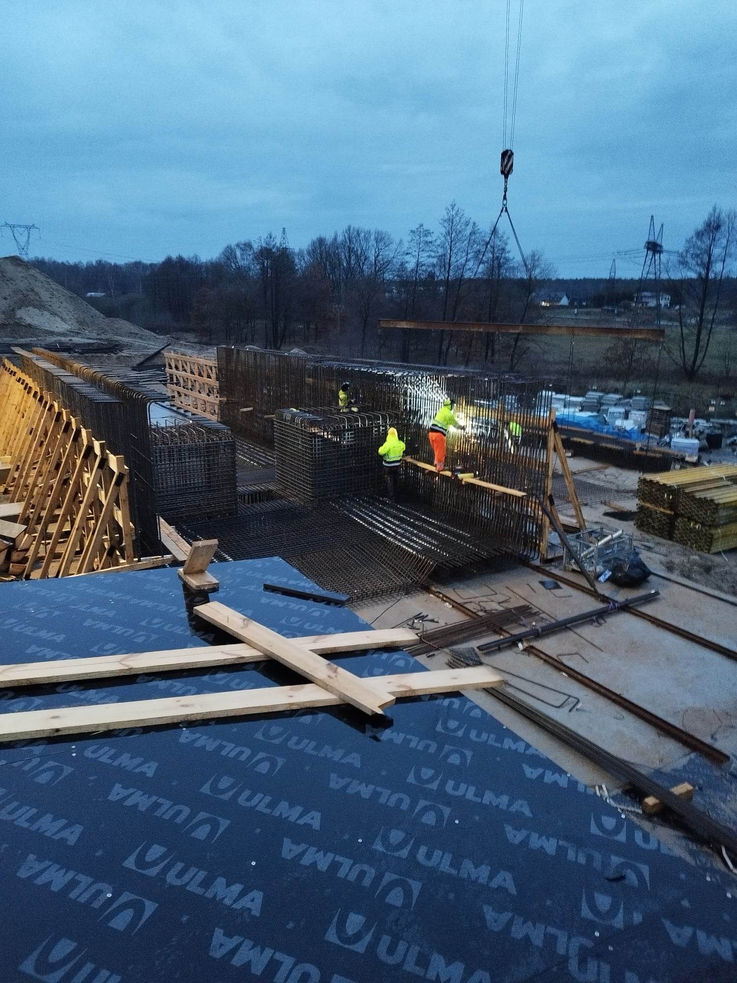 Konstrukcja żelbetowa w trakcie budowy: pracownicy w odblaskowych ubraniach montują zbrojenie na tle pochmurnego nieba, dźwig podnosi stalowy element, w tle widoczne elementy szalunków i materiały...