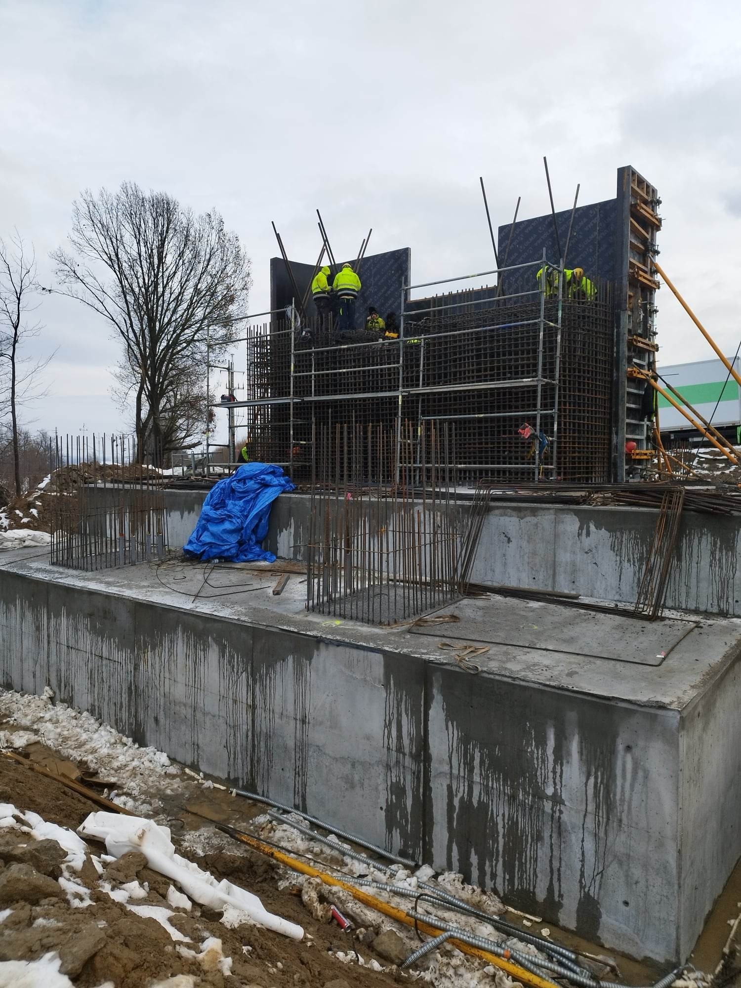Betonowa konstrukcja w trakcie budowy z widocznym zbrojeniem i deskowaniem, pracownicy w żółtych kamizelkach na górze konstrukcji, niebieska plandeka i resztki śniegu w otoczeniu.