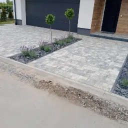 Nowoczesny podjazd z szarej kostki brukowej ułożonej we wzór, z obrzeżem z drobnych kamieni i nasadzeniami lawendy oraz dwoma kulistymi drzewkami, na tle domu z garażem i drewnianymi elementami...