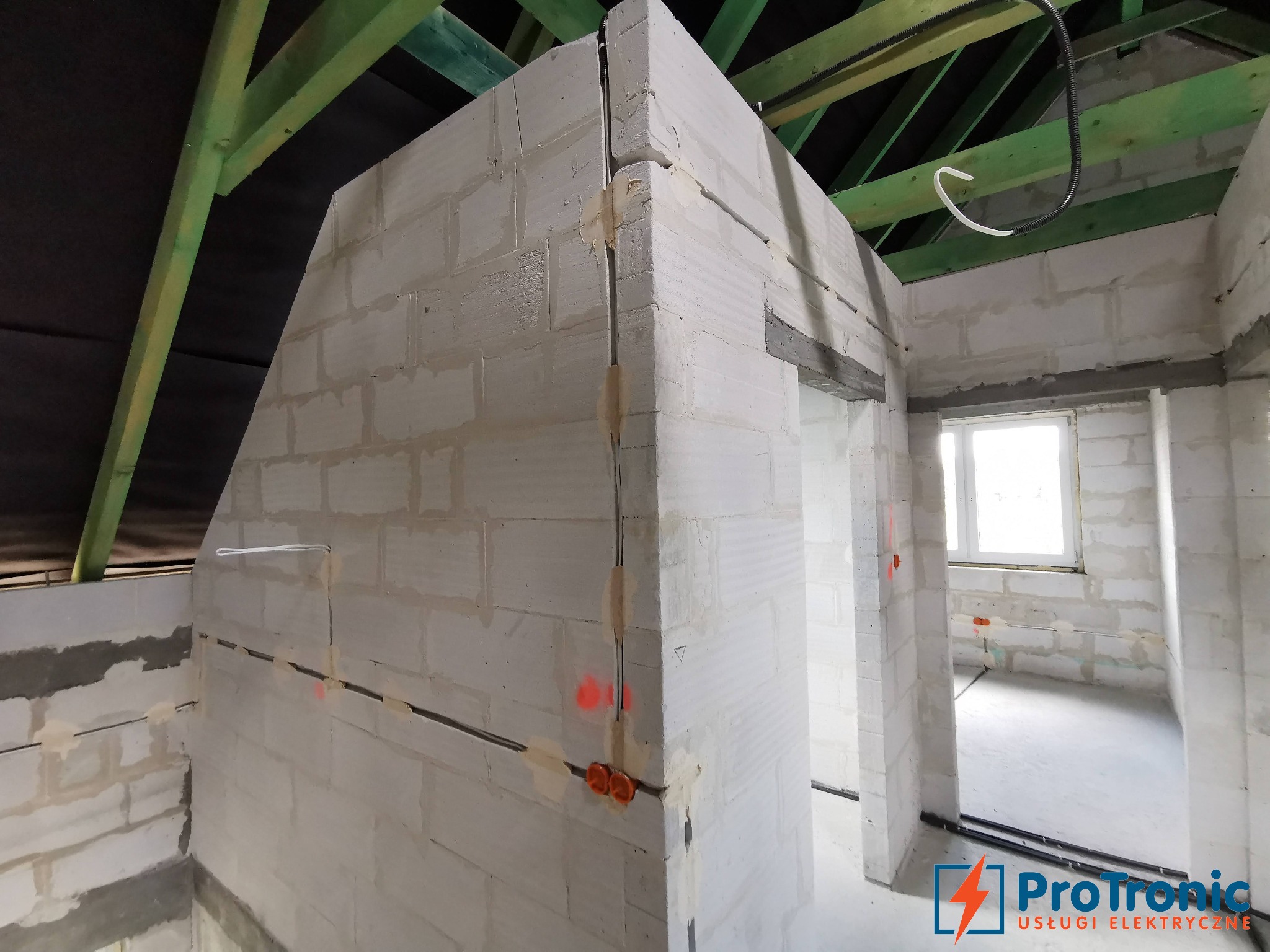 Wnętrze w trakcie budowy z widoczną instalacją elektryczną w bruzdach ścian z betonu komórkowego, zielona więźba dachowa i logo firmy elektrycznej w prawym dolnym rogu.