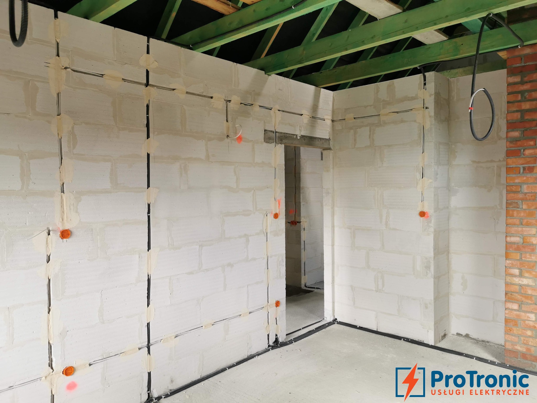 Wnętrze w trakcie prac elektrycznych: widoczne przewody poprowadzone po ścianach z betonu komórkowego, zabezpieczone gipsem, z pomarańczowymi puszkami instalacyjnymi, oraz logo firmy elektrycznej...