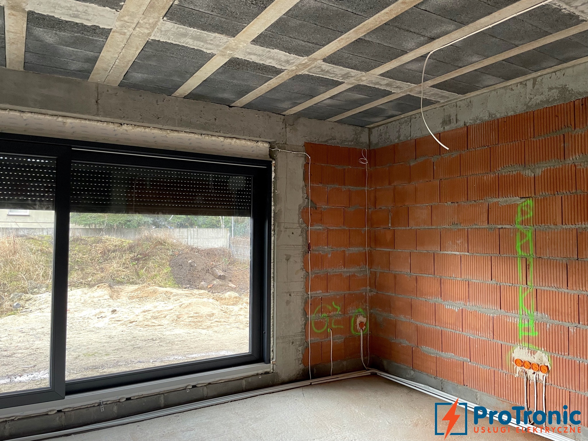 Surowy stan wnętrza w Krosnie z widocznymi przewodami elektrycznymi na ścianie z cegły, instalacja podtynkowa, okno z roletami zewnętrznymi i betonowy sufit.