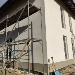 Dwukolorowa elewacja domu jednorodzinnego w trakcie ocieplania, widoczne rusztowanie, drabina oparta o ścianę, oraz narzędzia pracy na ziemi.