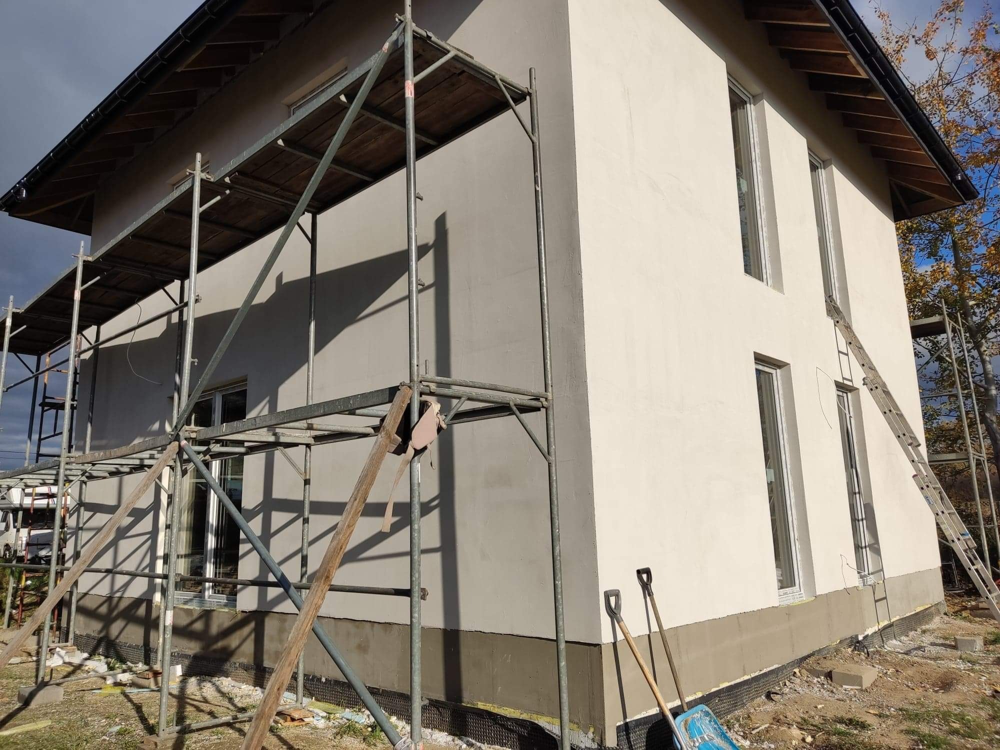 Dwukolorowa elewacja domu jednorodzinnego w trakcie ocieplania, widoczne rusztowanie, drabina oparta o ścianę, oraz narzędzia pracy na ziemi.