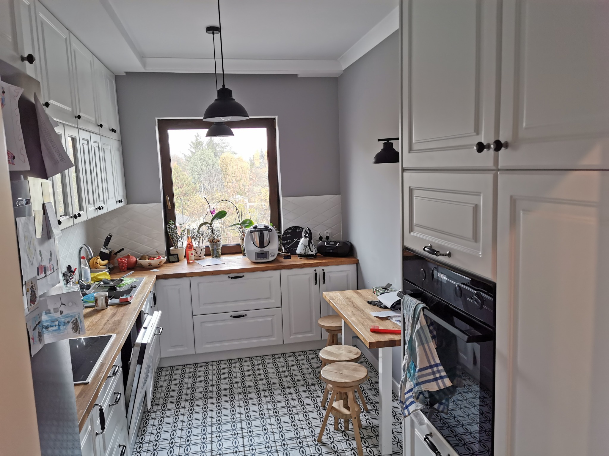 Jasna kuchnia w stylu skandynawskim z białymi szafkami, drewnianymi blatami i wzorzystą podłogą, oświetlona czarnymi lampami wiszącymi i kinkietem.