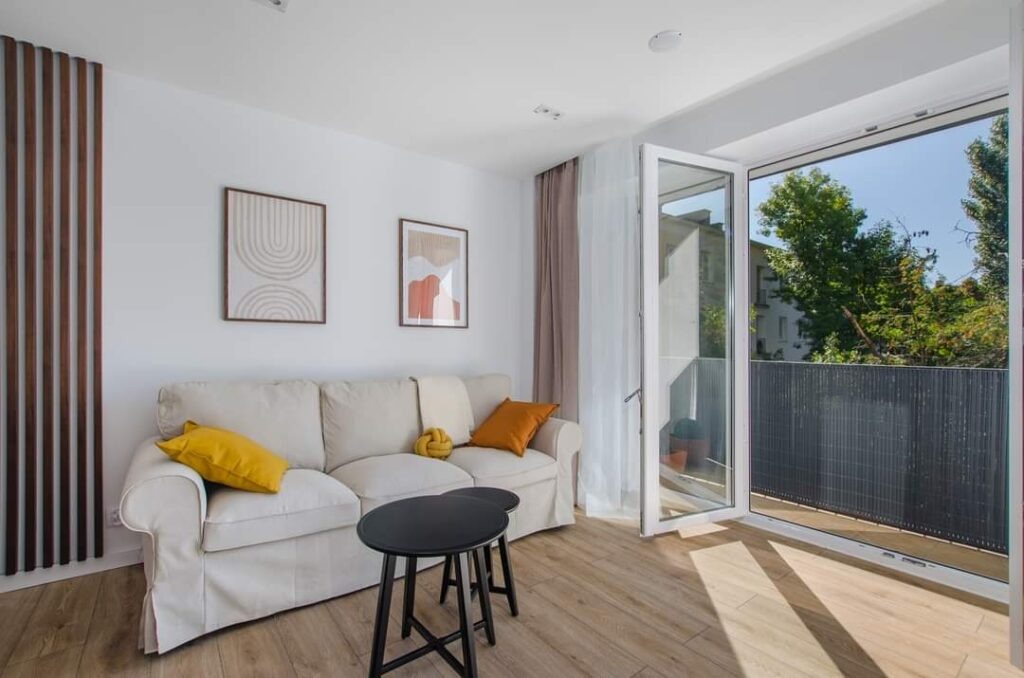 Jasny salon z drewnianą ścianką działową, beżową sofą z kolorowymi poduszkami, czarnymi stolikami kawowymi i otwartym balkonem z widokiem na zieleń.