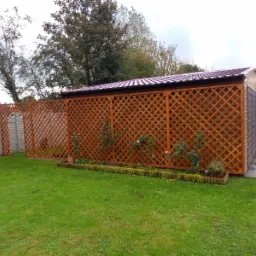 Drewniana altana z kratownicą i dachem z blachy trapezowej, obok trawnik, kwiaty i betonowe ogrodzenie.