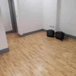 Jasna, drewniana podłoga z panelami ułożonymi w jodełkę, szare listwy przypodłogowe, białe ściany z gniazdkami elektrycznymi i dwa stare telewizory kineskopowe.