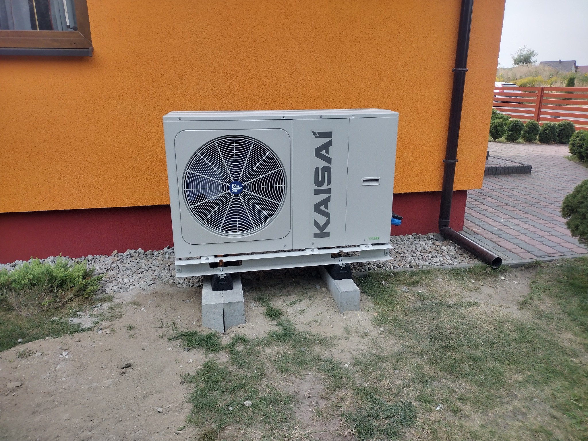 Zewnętrzna jednostka klimatyzacji Kaisai zamontowana na betonowych bloczkach przy pomarańczowej ścianie budynku z widoczną rynną spustową.