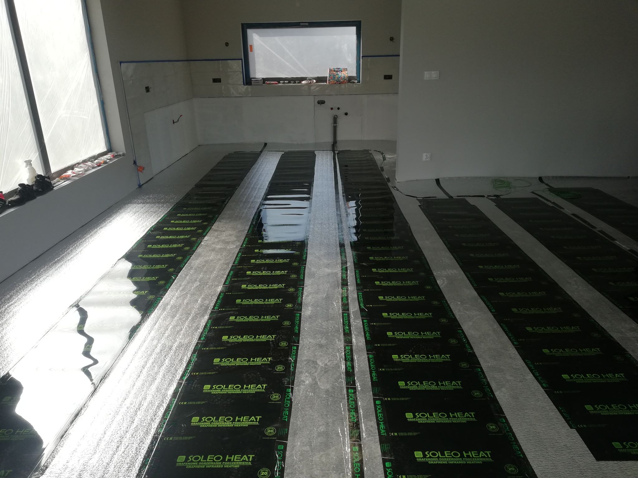 Instalacja folii grzewczej pod podłogę w nowym pomieszczeniu, widoczne paski folii SOLEO HEAT na srebrnej izolacji, ściany przygotowane do wykończenia.