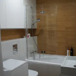 Nowoczesna łazienka z wanną za szklanym parawanem, umywalką nablatową i podwieszaną toaletą, wykończona płytkami imitującymi drewno.