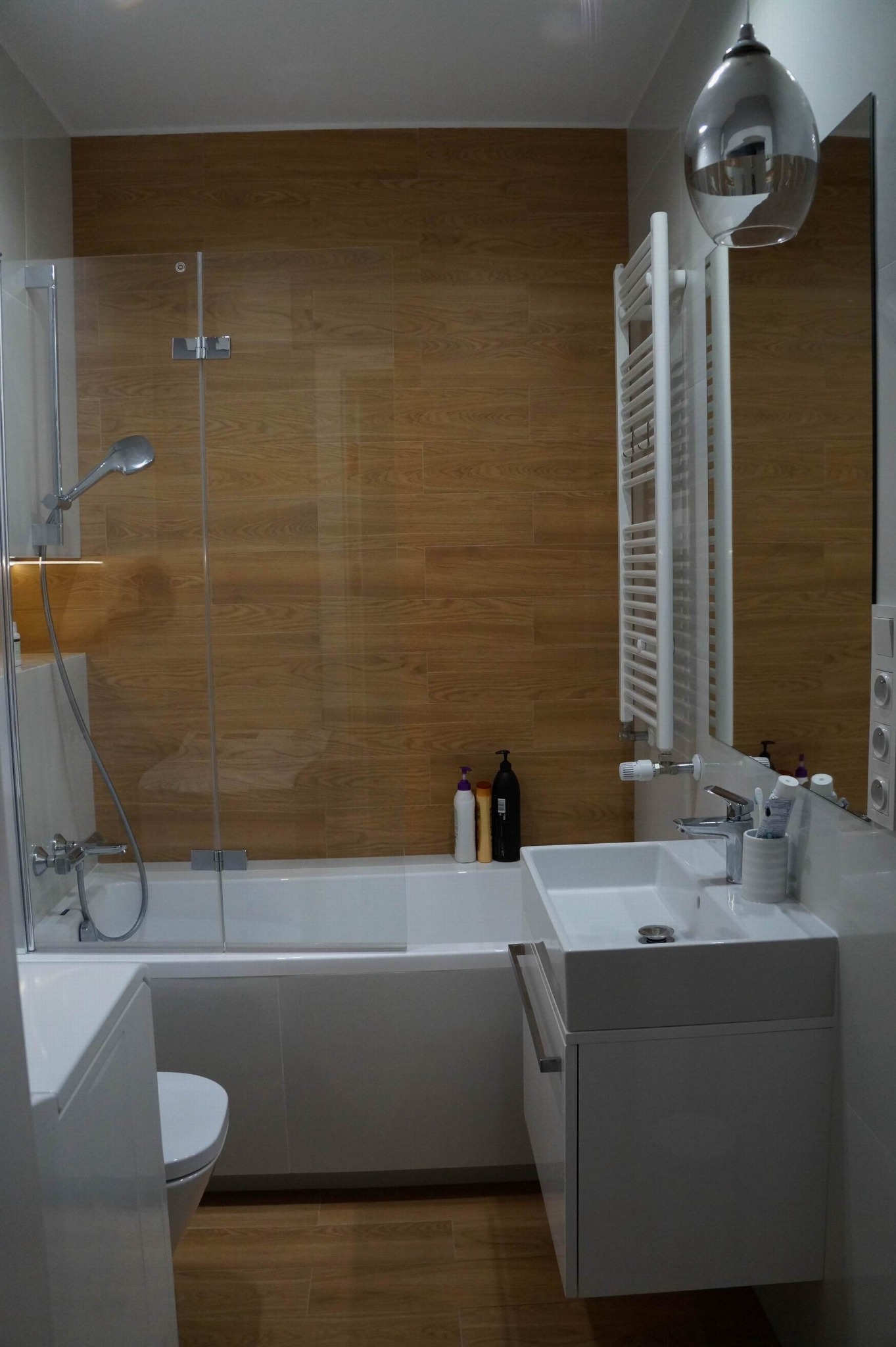 Nowoczesna łazienka z wanną za szklanym parawanem, umywalką na szafce i grzejnikiem drabinkowym, wykończona płytkami imitującymi drewno.