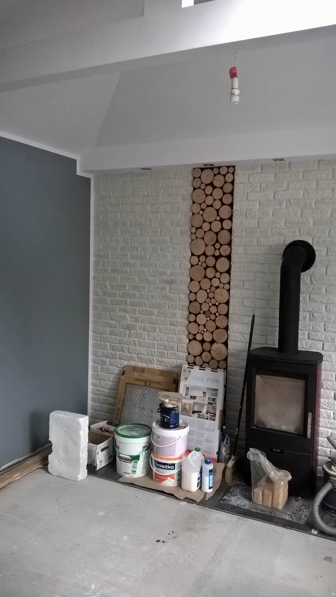 Wnętrze w trakcie remontu: ściana z cegły z dekoracyjnym panelem z drewna, obok piec wolnostojący, widoczne farby, wiadra i narzędzia malarskie, surowy beton na podłodze, wisząca żarówka.