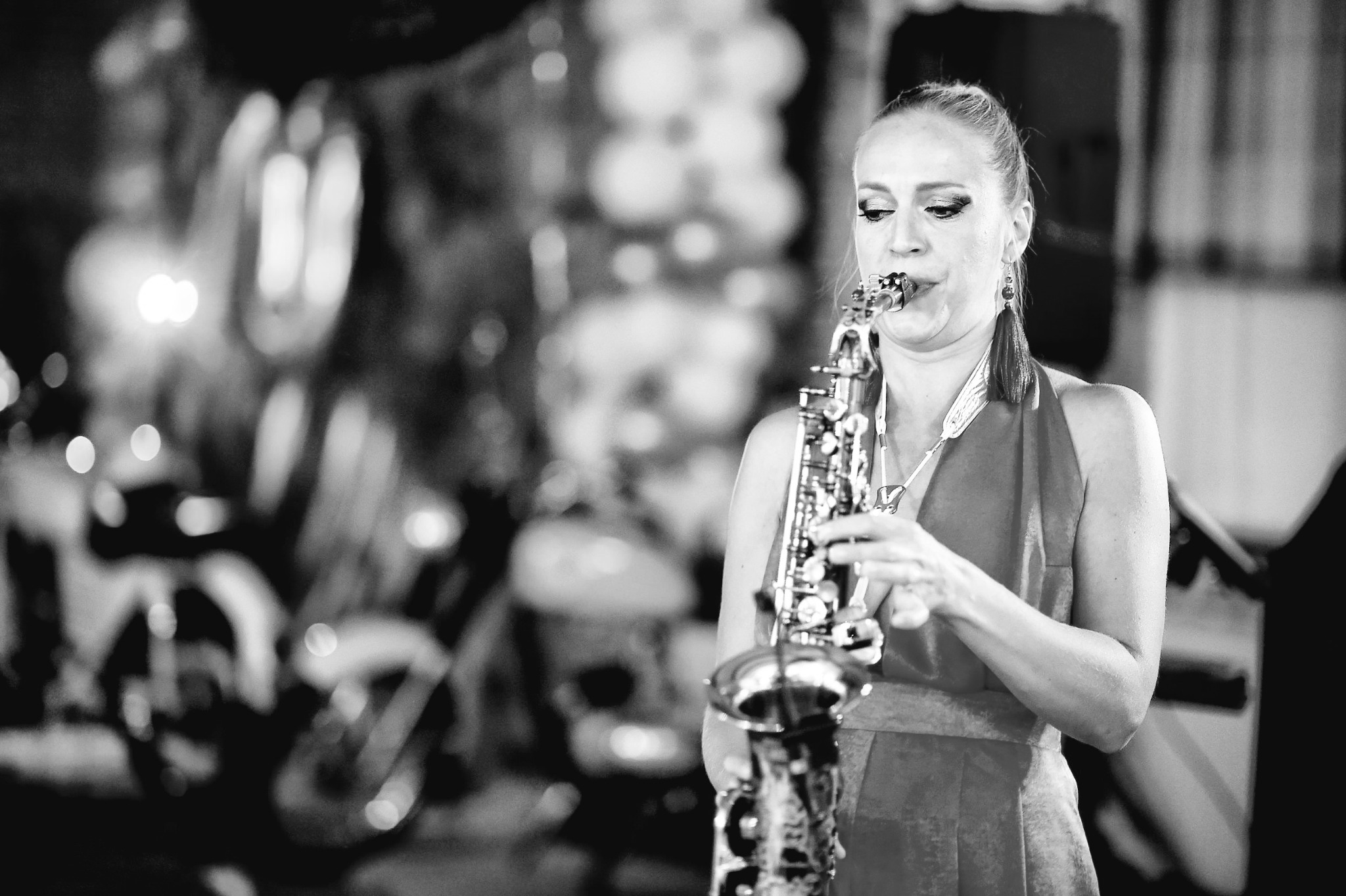 Saksofonistka w Gdyni gra na saksofonie tenorowym w plenerze, w tle rozmyte motocykle i oświetlenie sceniczne, zdjęcie czarno-białe.