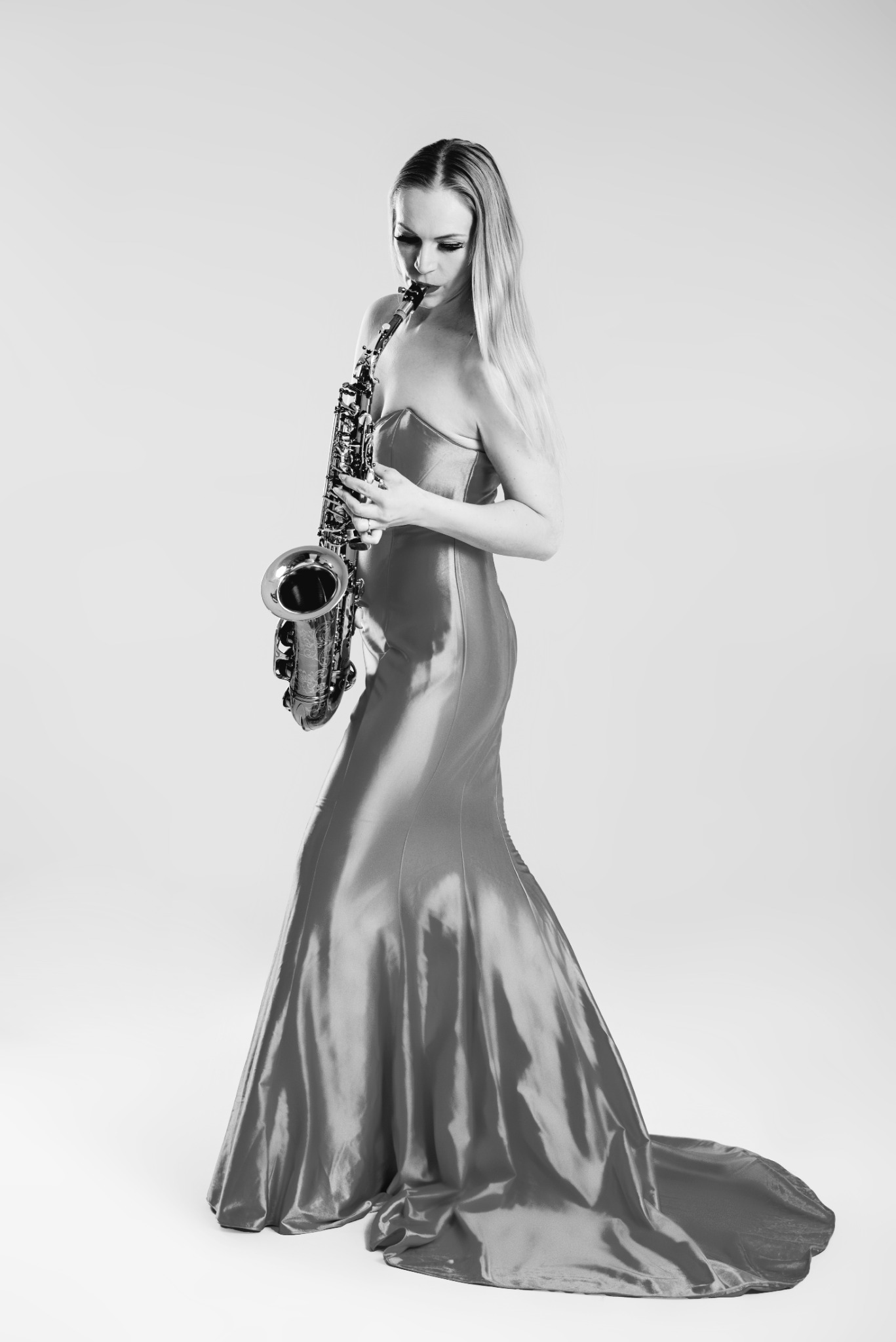 Kobieta w długiej, błyszczącej sukni gra na saksofonie w studio na jasnym tle. Ujęcie z profilu, skupione na instrumencie i twarzy muzyka.