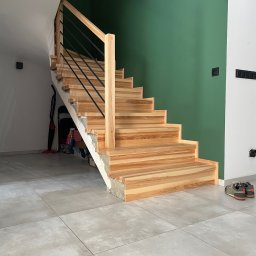 Adam Jendruś - Jasne, drewniane schody z czarnymi, metalowymi prętami balustrady, na tle ściany w kolorze butelkowej zieleni. Widoczne buty na podłodze i przedmioty pod schodami.