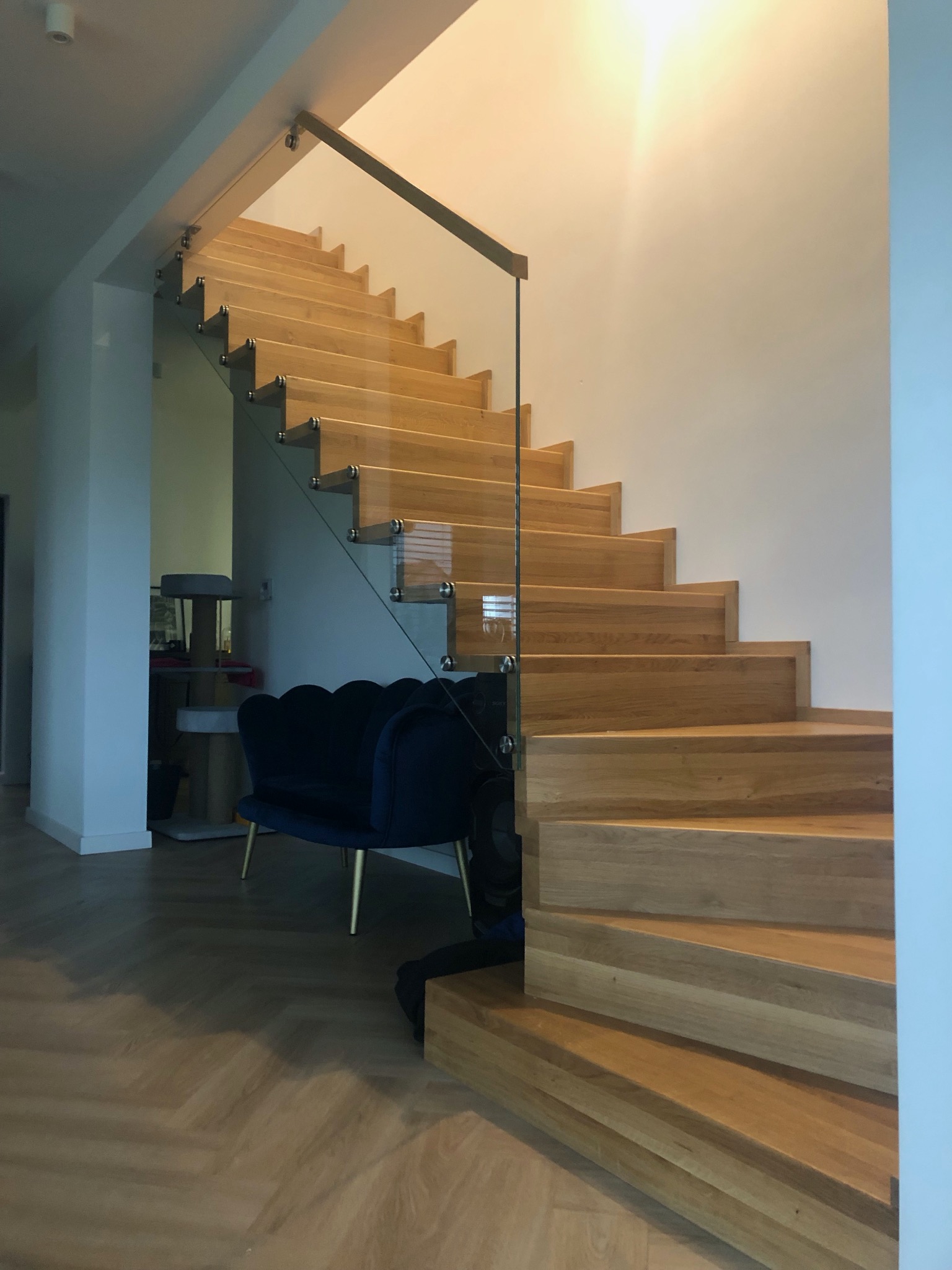 Jasne, dębowe stopnie schodów z przezroczystą szklaną balustradą, zamocowaną metalowymi uchwytami, w nowoczesnym wnętrzu z parkietem w jodełkę i niebieskim fotelem.